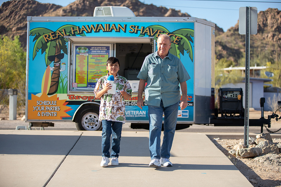 A man and a boy holding a blue sno cone walk away from a shaved ice truck