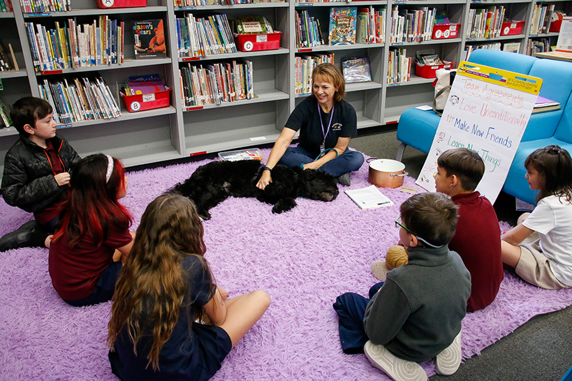 Students sit in a circle on a purple fuzzy carpet, with a woman and a fluffy black therapy dog in the center
