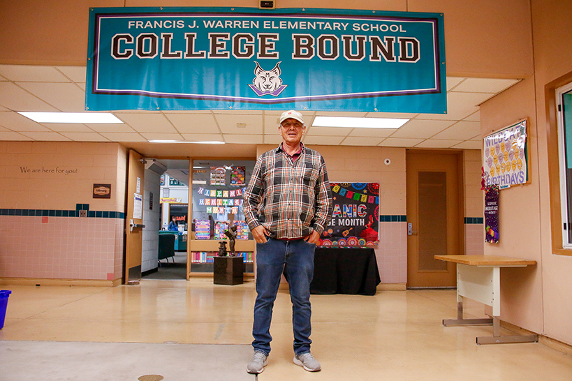 A man in a plaid shirt and jeans stands in the hallway underneath a banner reading Frances J. Warren Elementary School College Bound