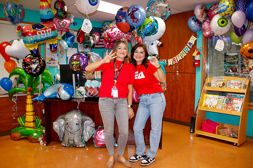 Two women in red shirts and jeans pose holding up the number 40 in front of balloons