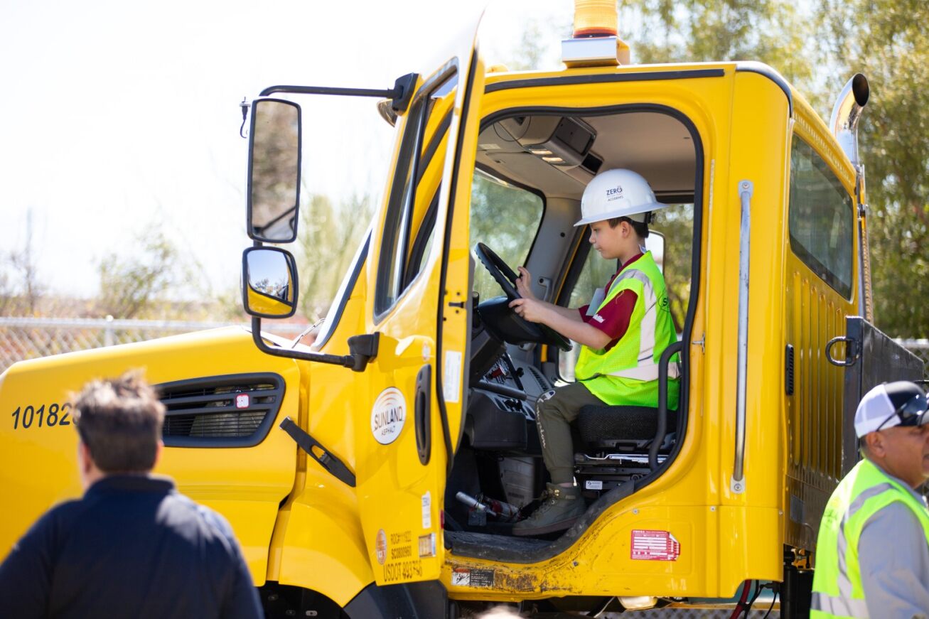 A boy in a white hard hat sits behind the wheel of a yellow asphalt truck