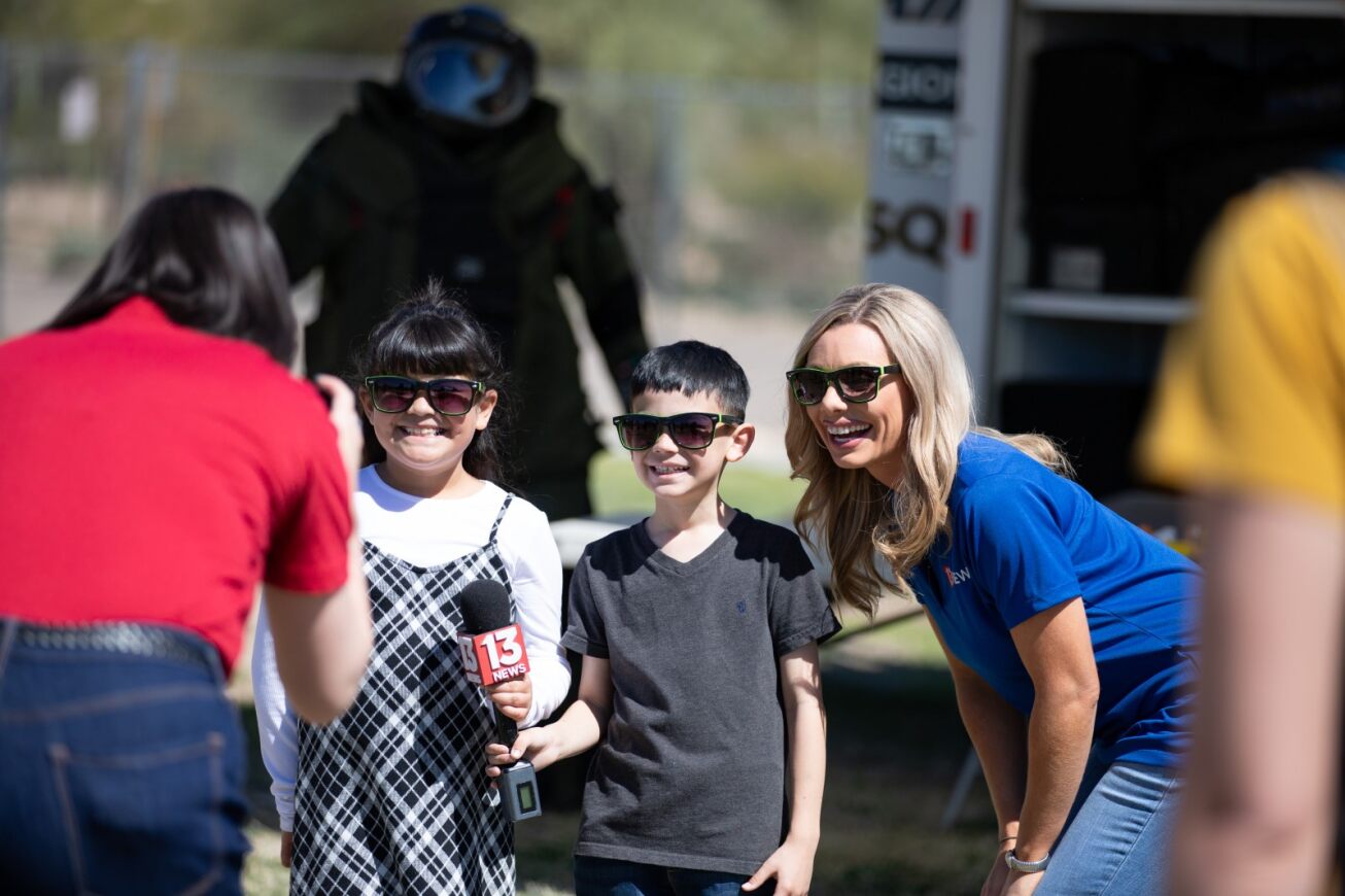 A girl and a boy wearing sunglasses smile next to a reporter from 13 News.